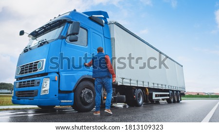 Truck Driver Crosses the Road in the Rural Area and Gets into His Blue Long Haul Semi-Truck with Cargo Trailer Attached. Logistics Company Moving Goods Across Countrie and Continent Royalty-Free Stock Photo #1813109323