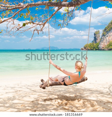Lady swinging on the picture perfect tropical beach with the view of the island and turquoise coral reef on a sunny summer day.