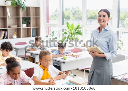 teacher looking at camera while holding book near multiethnic pupils writing during lesson