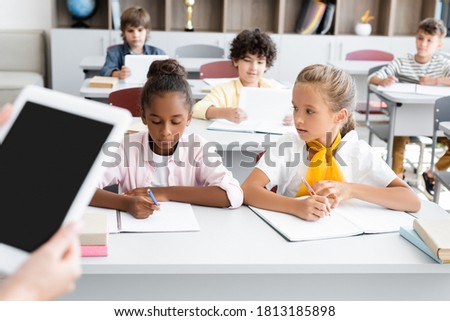 cropped view of teacher holding digital tablet with blank screen near multicultural pupils writing in notebooks