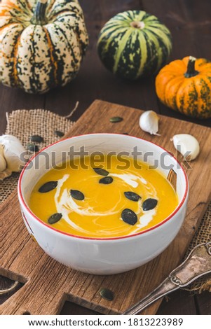 Bowl with pumpkin soup on a wooden board on dark brown rustic wooden background, decorated with little pumpkins and seeds. Vertical stock photo.