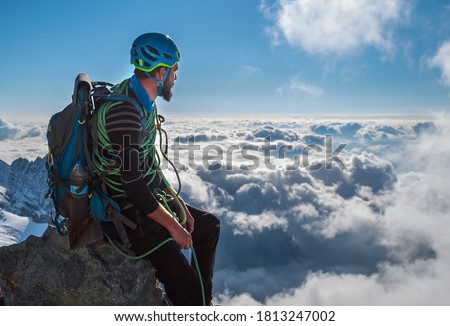Bearded Climber in a safety harness, helmet, and on body wrapped climbing rope with sitting at 3600m altitude on a cliff and looking at  picturesque clouds during Mont Blanc ascending, France route Royalty-Free Stock Photo #1813247002