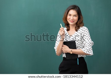 asian teacher holding chalk and book on green chalkboard background Royalty-Free Stock Photo #1813344112