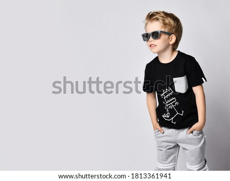 Cool blond kid boy in sunglasses, black t-shirt with dinosaur print and gray pants stands side to camera with hands in pockets looking aside at free copy space over gray background