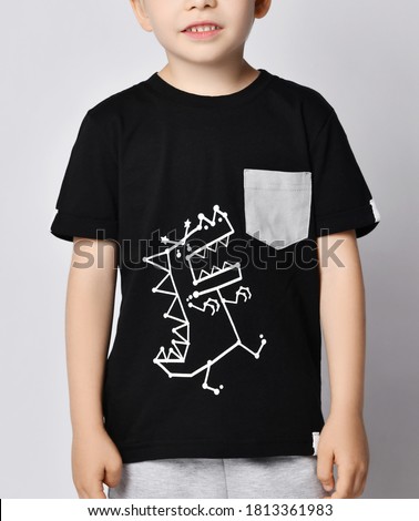 Closeup of blond kid boy in black t-shirt with roaring cartoon dinosaur print picture and gray pocket over gray background