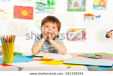 Nice bored three years old boy sitting in the class with images paper and pencils
