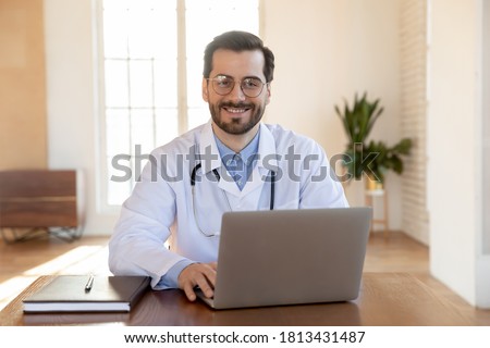 Head shot portrait smiling doctor wearing glasses working on laptop, sitting at desk in office, therapist physician gp wearing white medical uniform with stethoscope looking at camera