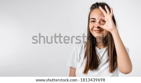 Close up portrait of carefree modern girl with long chestnut hairs, looking at the camera through fingers in OKAY gesture. Face expressions, emotions, and body language. Copy space for your text