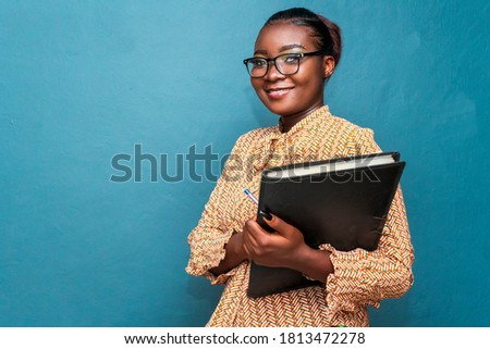 Bold,beautiful and confident African business woman or teacher holding a black file,isolated on blue studio background Royalty-Free Stock Photo #1813472278