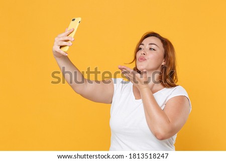 Pretty young redhead plus size body positive woman 20s in white casual t-shirt doing selfie shot on mobile phone blowing sending air kiss isolated on yellow color background studio portrait