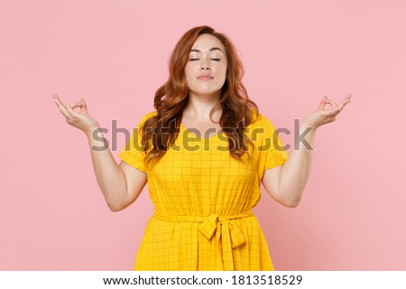 Young redhead plus size body positive woman 20s in yellow dress hold hands in yoga gesture, relaxing meditating, trying to calm down isolated on pastel pink color background studio portrait