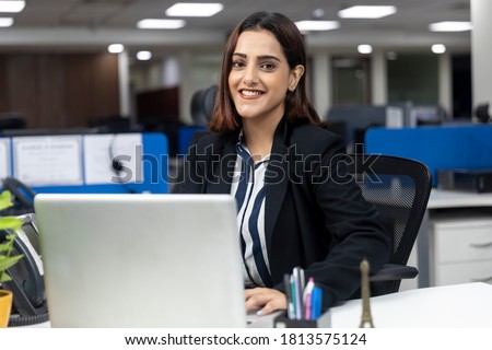Portrait of smiling pretty Indian businesswoman sitting at her workstation against office background, working on laptop, corporate environment, Royalty-Free Stock Photo #1813575124