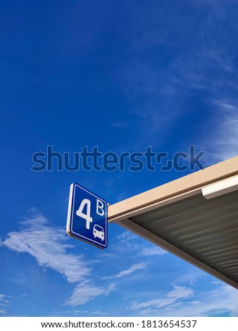A square metallic sign showing number and area zone in a parking lot. A sign hanging at a parking roof against blue sky and white clouds.
