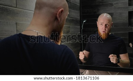A young, Caucasian, charismatic, bald guy with a red beard invigorates himself in front of the mirror, talking to his reflection in a modern bathroom. Morning mood for the day. Recharge with energy. Royalty-Free Stock Photo #1813671106