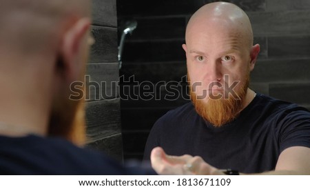 A young, Caucasian, charismatic, bald guy with a red beard with a serious look pokes his finger at his reflection in mirror, talking and recharging himself with energy.Modern bathroom. Morning rituals Royalty-Free Stock Photo #1813671109