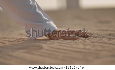 Closeup of the hand of a Caucasian man in white clothing. He runs his hand over the sand, takes it into a fist, and then pours it back, turning it over in his hand. Side view. Sunny weather. Royalty-Free Stock Photo #1813673464