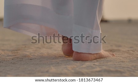 Caucasian man in white long clothes to the heel, walking along the sand with slow steps, taking his feet off the ground, you can see the feet. Slow step. Back view. Close-up. Royalty-Free Stock Photo #1813673467