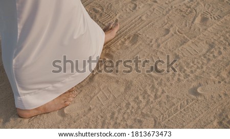 Caucasian man in white, long clothes up to the heels walking along the sand in small steps. Bare feet walking along the sand. On the sand other footprints from the wheels and wheels of cars. Royalty-Free Stock Photo #1813673473