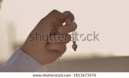 A man looks at a chain with a pendant, inside of which is a pearl. The chain is in his hand, squeezed into a fist. Pendant swaying in the wind. Warm tones. Close-up. Royalty-Free Stock Photo #1813673476