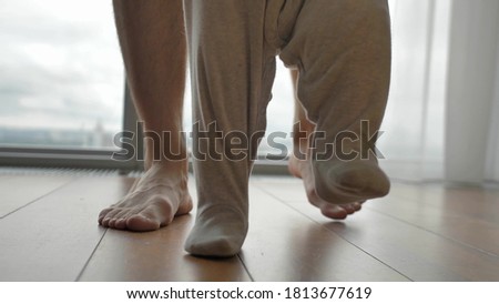 A little baby in a gray jumpsuit makes his first steps with the support of his dad. Close-up of baby and father's feet. Bright room, parquet floor, windows to the floor. Royalty-Free Stock Photo #1813677619