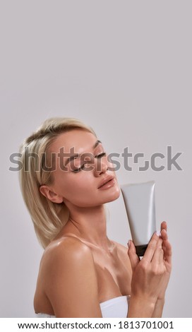 Portrait of young woman with perfect fresh skin holding cosmetic skincare, bodycare product while posing isolated over grey background, Vertical shot