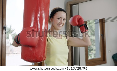 The woman is boxing at home. Woman with boxing gloves looking at camera and holding punching bag. Strong woman portrait.She smiles at the camera.