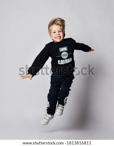 Active frolic blond kid boy in black jersey sweater with printed words inscription on it jumps high over gray background. Translation: "Want. Can. Will Do"