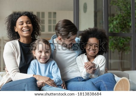 Happy emotional mixed race family relaxing together on comfortable couch indoors. Laughing multiracial couple enjoying watching television funny films with joyful small biracial children at home. Royalty-Free Stock Photo #1813838684