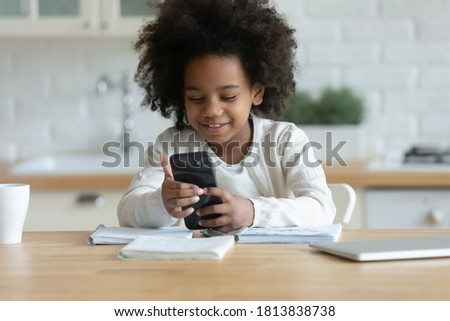 Head shot adorable smiling little mixed race kid girl sitting at table, using smartphone applications alone at home. Happy small african ethnicity biracial child involved in mobile game or web surfing
