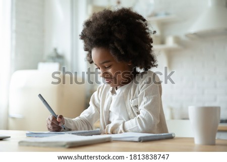 Smiling adorable beautiful african ethnicity little girl sitting at table, writing notes in copybook. Happy small school aged mixed race kid involved in preparing interesting homework alone indoors. Royalty-Free Stock Photo #1813838747