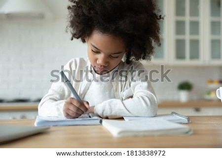 Head shot focused small 7 years old african american child girl sitting at table, handwriting school tasks alone at home, serious busy little mixed race female pupil studying remotely, education Royalty-Free Stock Photo #1813838972
