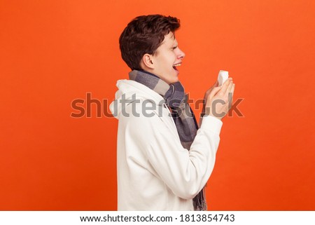 Profile of upset ill teenager in white casual sweatshirt and with checkered scarf on neck widely open mouth sneezing into paper napkin, cold. Indoor studio shot isolated on orange background