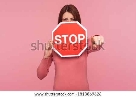 Angry strict woman in pink sweater holding stop warning sign and pointing finger at you, access forbidden, prohibition. Indoor studio shot isolated on pink background