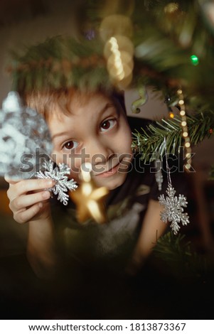 Little cute caucasian boy peeping out of Christmas tree with twinkling decorations. He is holding a snowflake. Image with selective focus