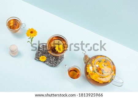 Cup of herbal tea and transparent teapot with calendula flowers on blue background. Calendula Tea Benefits Your Health concept. Top view Flat lay.