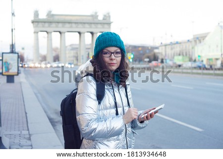 woman in a down jacket and hat stands at a public transport stop and uses a smartphone. Wait for the bus. Choose a route in the mobile app, search for a bus route or call a taxi on your phone