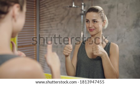 Blond haired young woman looking at her reflection in the mirror and smiling.Showing ok sign and thumbs up.