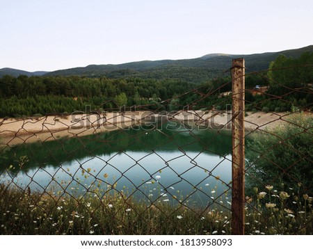 A forest area surrounded by an old wire fence and a green lake are seen. A very beautiful view of nature is in the photo.