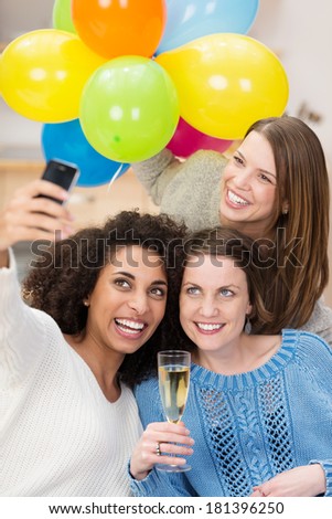 Group of friends celebrating with flutes of champagne taking a self portrait on a mobile phone with a bunch of colorful party balloons as they laugh and joke
