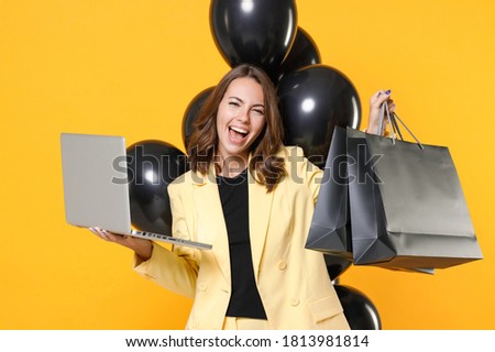 Joyful young woman 20s wearing suit jacket hold package bags with purchases after shopping bunch of air balloons laptop pc computer isolated on yellow background studio portrait. Black friday sale