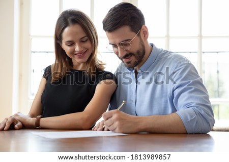 Excited young Caucasian couple sign contract buying first shared house together, overjoyed smiling husband and wife sit at desk put signature on document agreement closing deal at office meeting