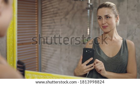Beautiful woman with cheerful blonde hair smiling and taking selfie on cell phone while looking in bathroom mirror.