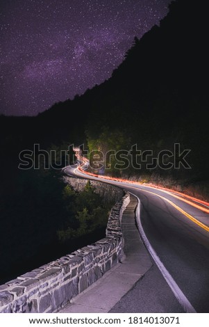 A long exposure of cars passing through a winding road on the side of a mountain under the milky way sky at night