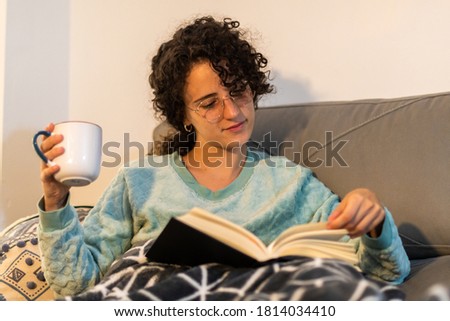 Stock photo of a beautiful and happy young woman with curly hair and glasses reading a book at home, sitting on the coach. Curly girl with soft blanket and winter pajama reading on a winter afternoon.
