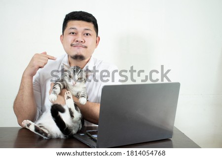 Young bearded businessman sits in desk and uses laptop, hugging a cute cat. In a white background. Man working, blogging.