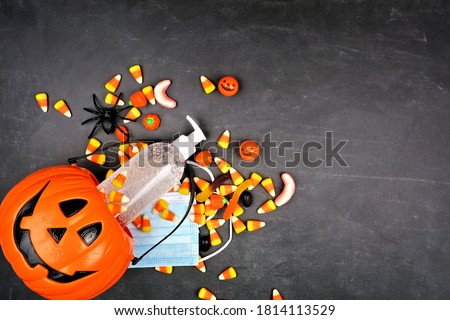 Halloween Jack o Lantern pail with spilling candy and coronavirus prevention supplies. Above view over a black background.