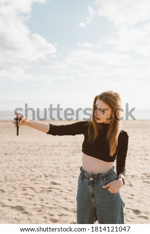 Pretty female with long hair, blonde takes photo on mobile phone on sandy beach in summer or autumn. Beautiful woman in jeans and black top