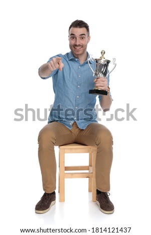 Positive smart casual man holding trophy and pointing forward while sitting on a chair on white studio background