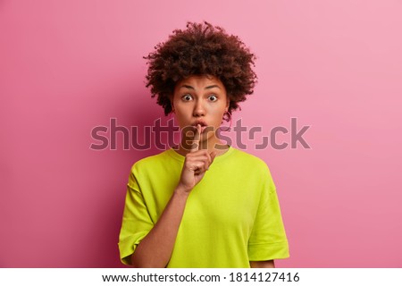 Surprised dark skinned woman makes silence gesture, keeps finger over lips and looks mysterious at camera. Wondered curly millennial girl shows hush sign, poses against rosy background. Body language