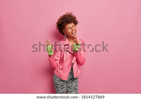 Indoor shot of glad joyful woman keeps hand near mouth as microphone, pretends singing loudly and wears stylish sport clothes. Brunette curly haired Afro American female sings favorite song.